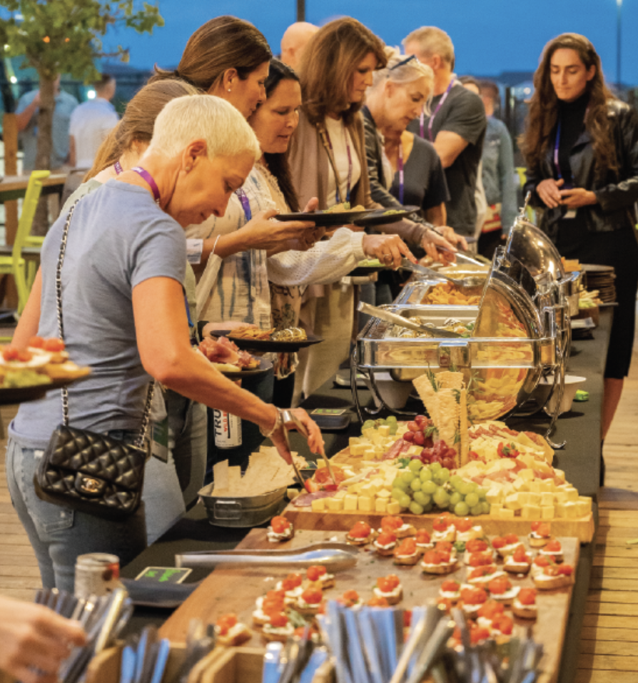 A group of people serve themselves food from a buffet table filled with cheese, fruit, and appetizers at an outdoor event in the evening.