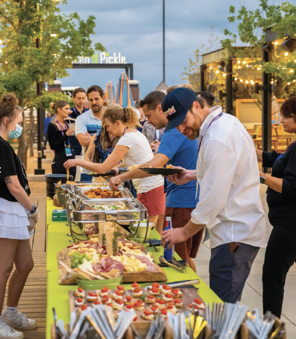People serve themselves food from a colorful outdoor buffet table at a lively gathering. String lights and trees decorate the area, and several people are smiling and chatting in the background.