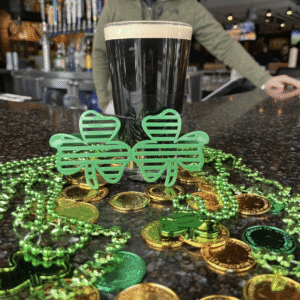 A pint of dark beer on a bar counter with green shamrock glasses, green bead necklaces, and gold coins, suggesting a St. Patrick’s Day celebration. A person stands in the blurred background.