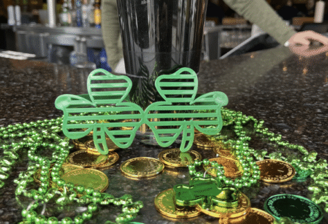 A pint of dark beer on a bar counter with green shamrock glasses, green bead necklaces, and gold coins, suggesting a St. Patrick’s Day celebration. A person stands in the blurred background.