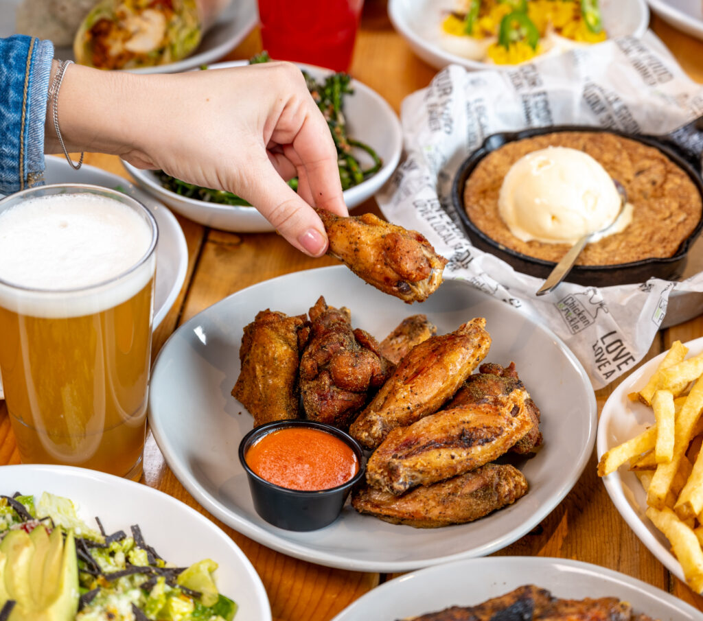 A hand holds a chicken wing above a plate of wings with dipping sauce. Surrounding dishes include fries, salad, a cookie with ice cream, and a glass of beer on a wooden table.