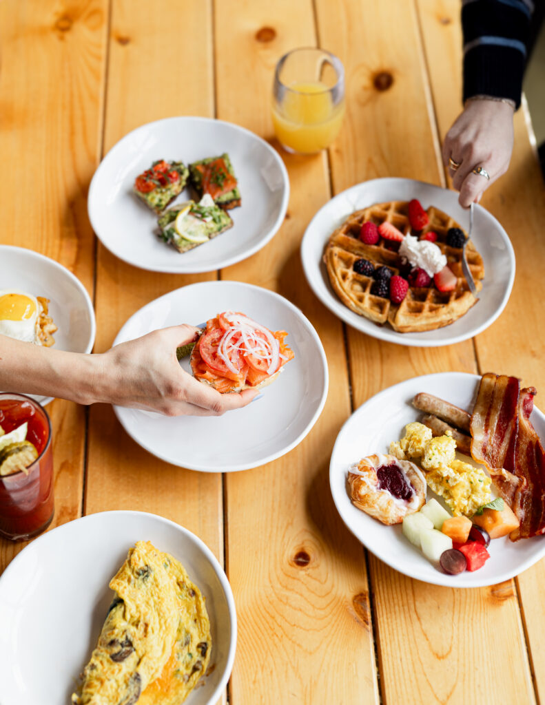 An overhead view of a wooden table with plates of breakfast foods, including waffles with berries, scrambled eggs, bacon, sausage, fruit, avocado toast, omelet, and drinks. Two hands are reaching for food.