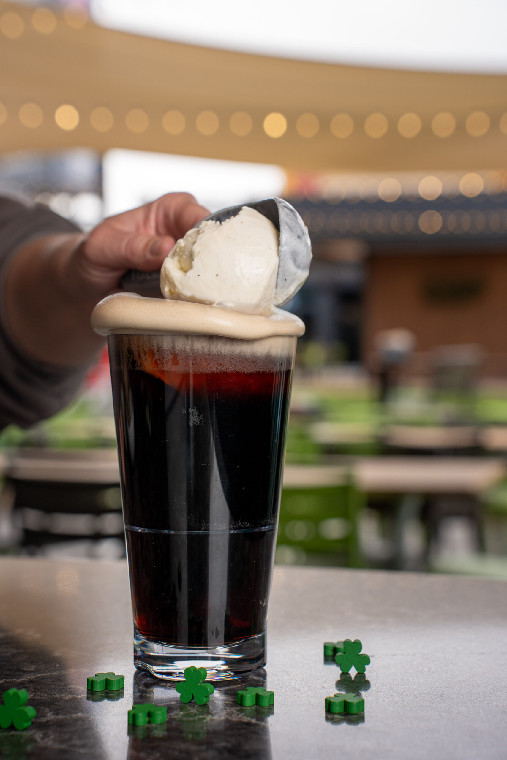 A hand places a scoop of vanilla ice cream on top of a pint of dark beer with foam, on a table decorated with small green shamrock shapes, with outdoor seating in the blurred background.