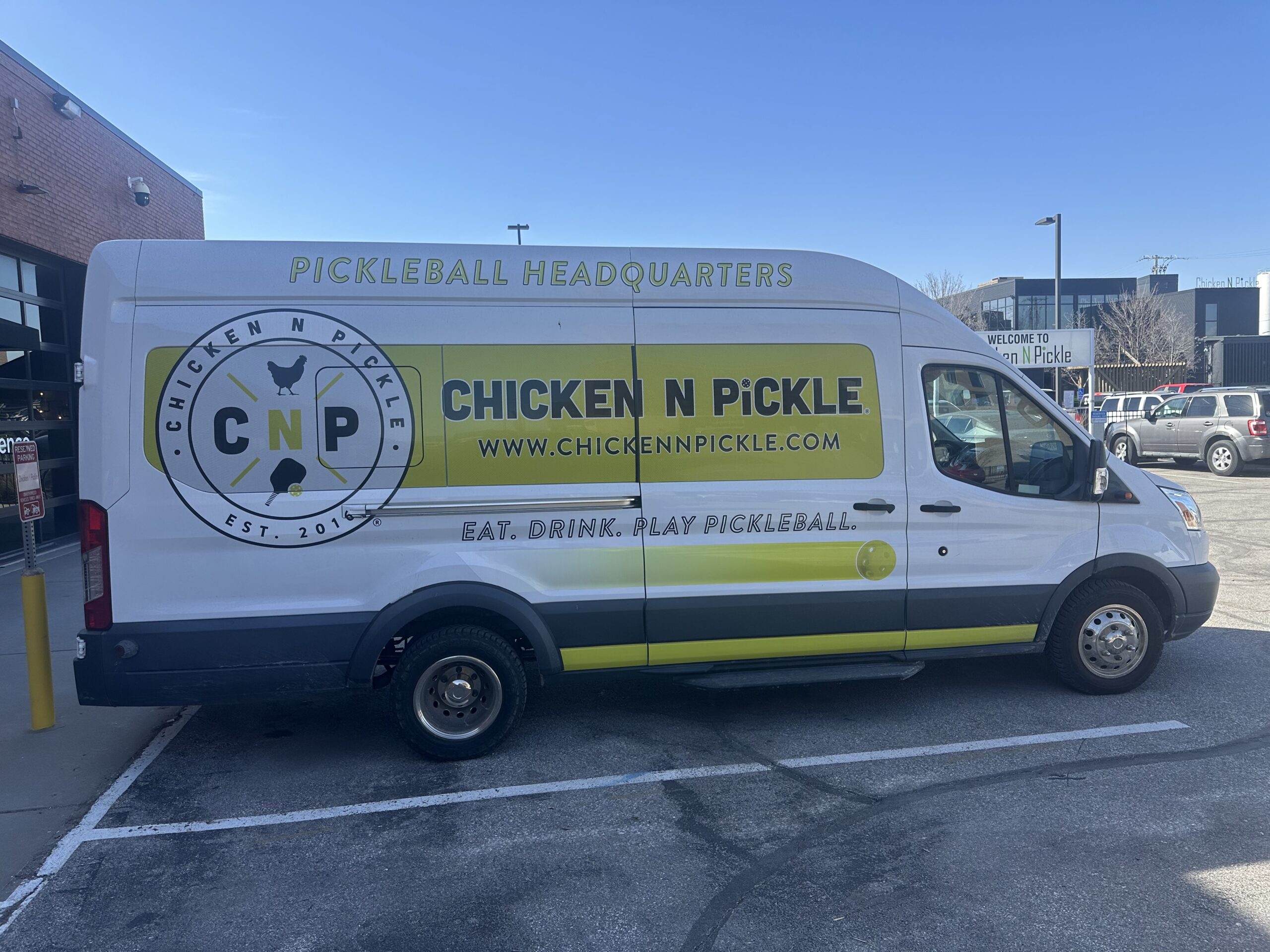 A white van parked in a lot features a large “Chicken N Pickle” logo and website, along with the phrases “Pickleball Headquarters” and “Eat. Drink. Play Pickleball” on its side.