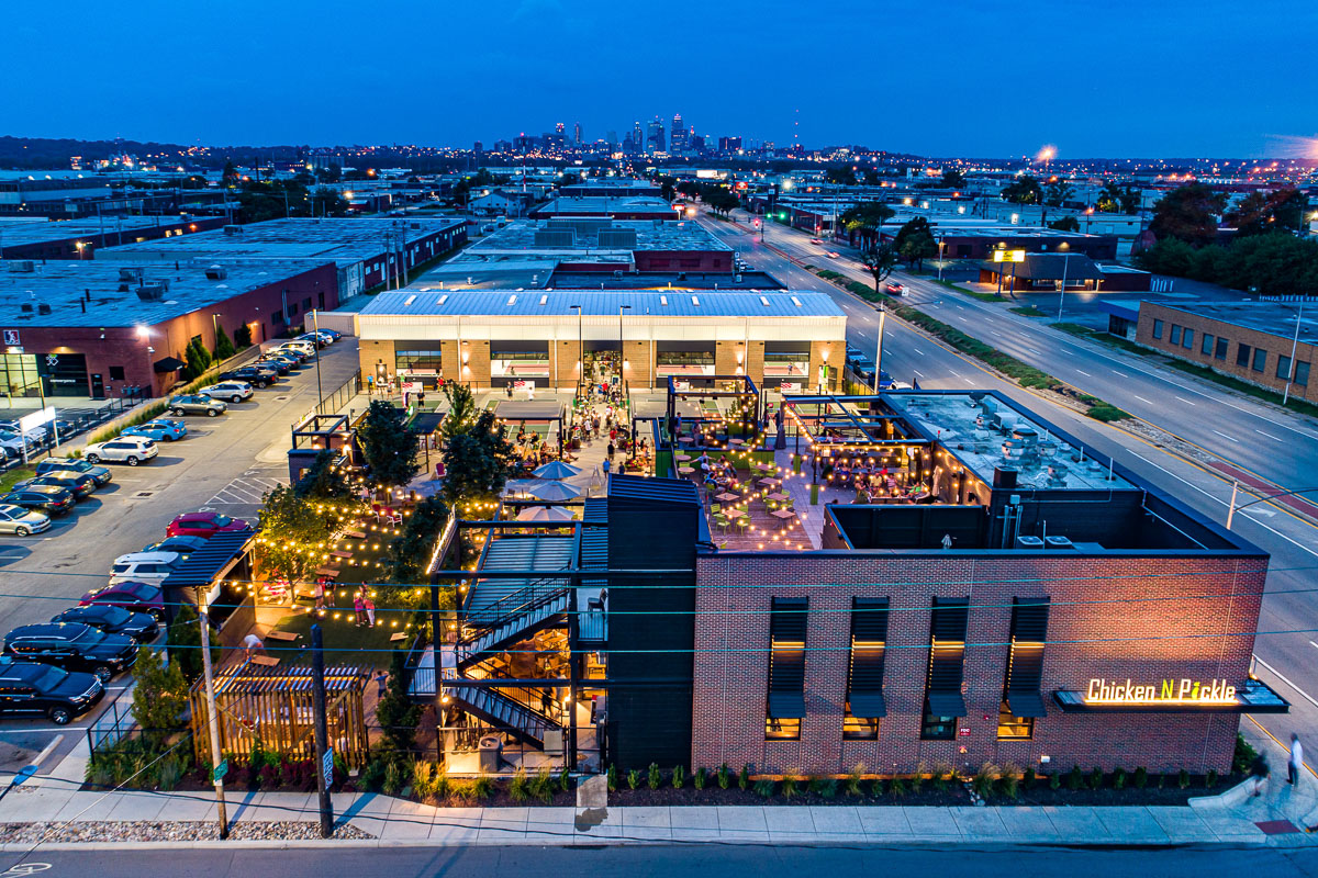 Aerial view of a lively, well-lit outdoor dining and activity area at Chicken N Pickle, surrounded by parking lots and industrial buildings, with a city skyline visible in the distance during dusk.