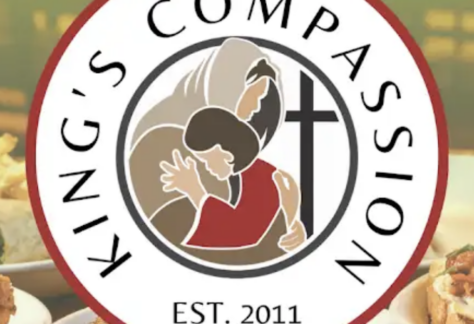 Circular logo for Kings Compassion featuring two people embracing, a large cross, and EST. 2011 text. Food dishes are blurred in the background, highlighting their giveback mission benefiting vets and youth in need.