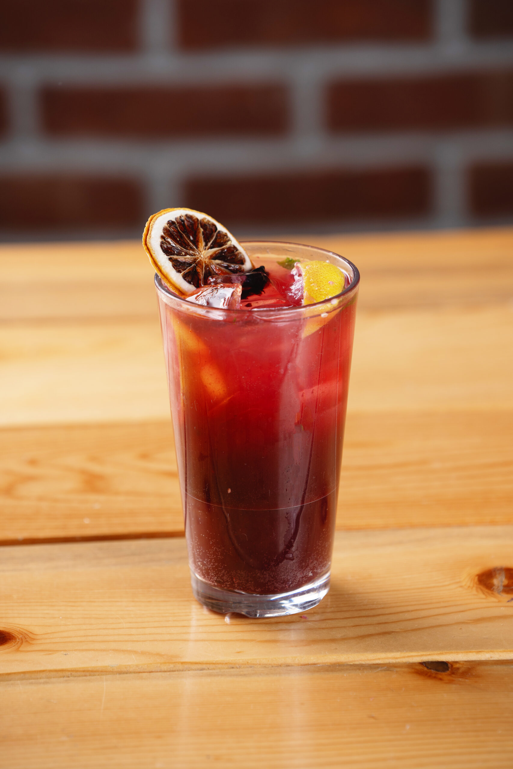 A glass of red cocktail with ice, garnished with a dried orange slice, sits on a wooden table with a blurred brick wall in the background.