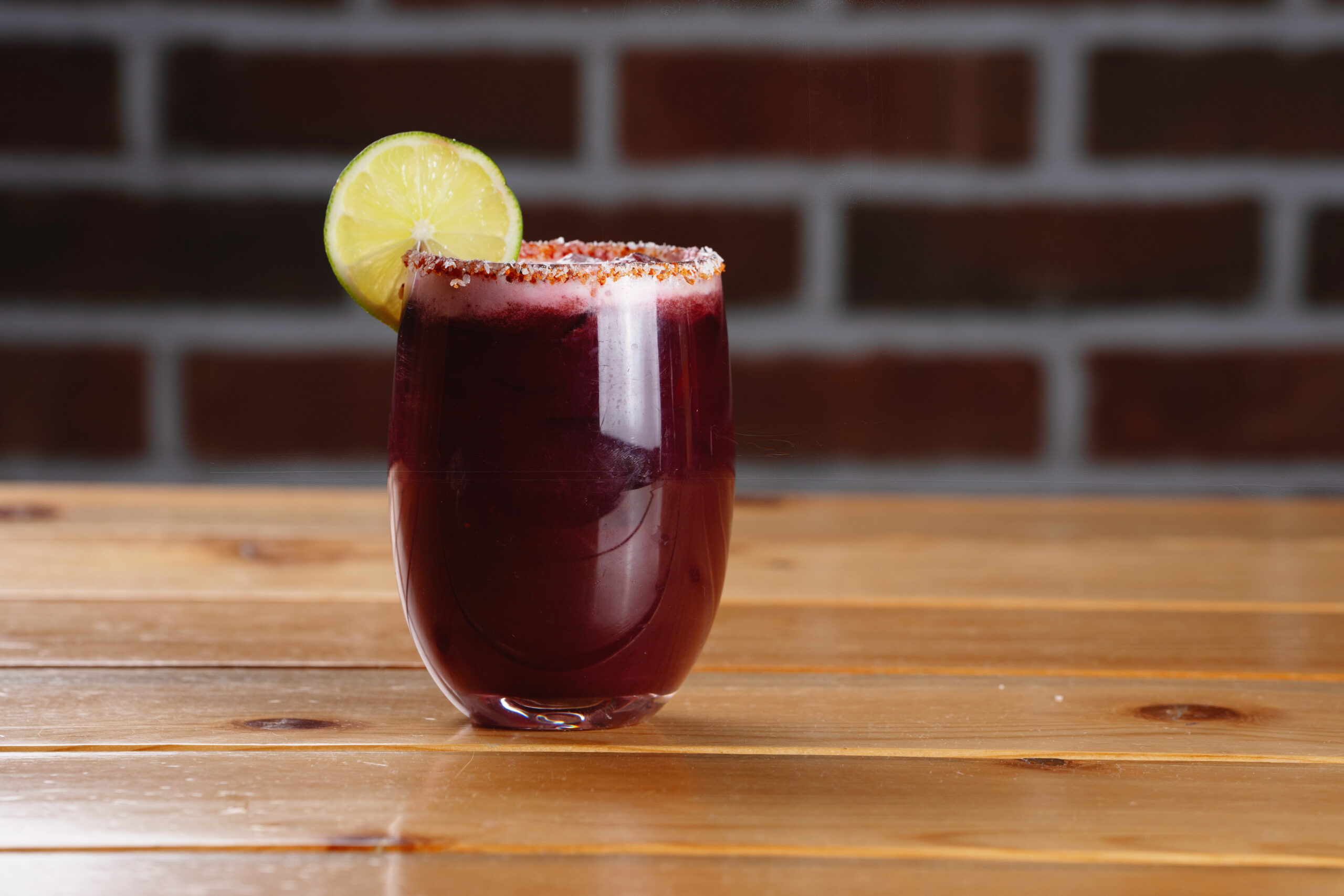 A glass of dark red beverage with a salted rim, garnished with a lime wheel, sits on a wooden table against a blurred brick wall background.