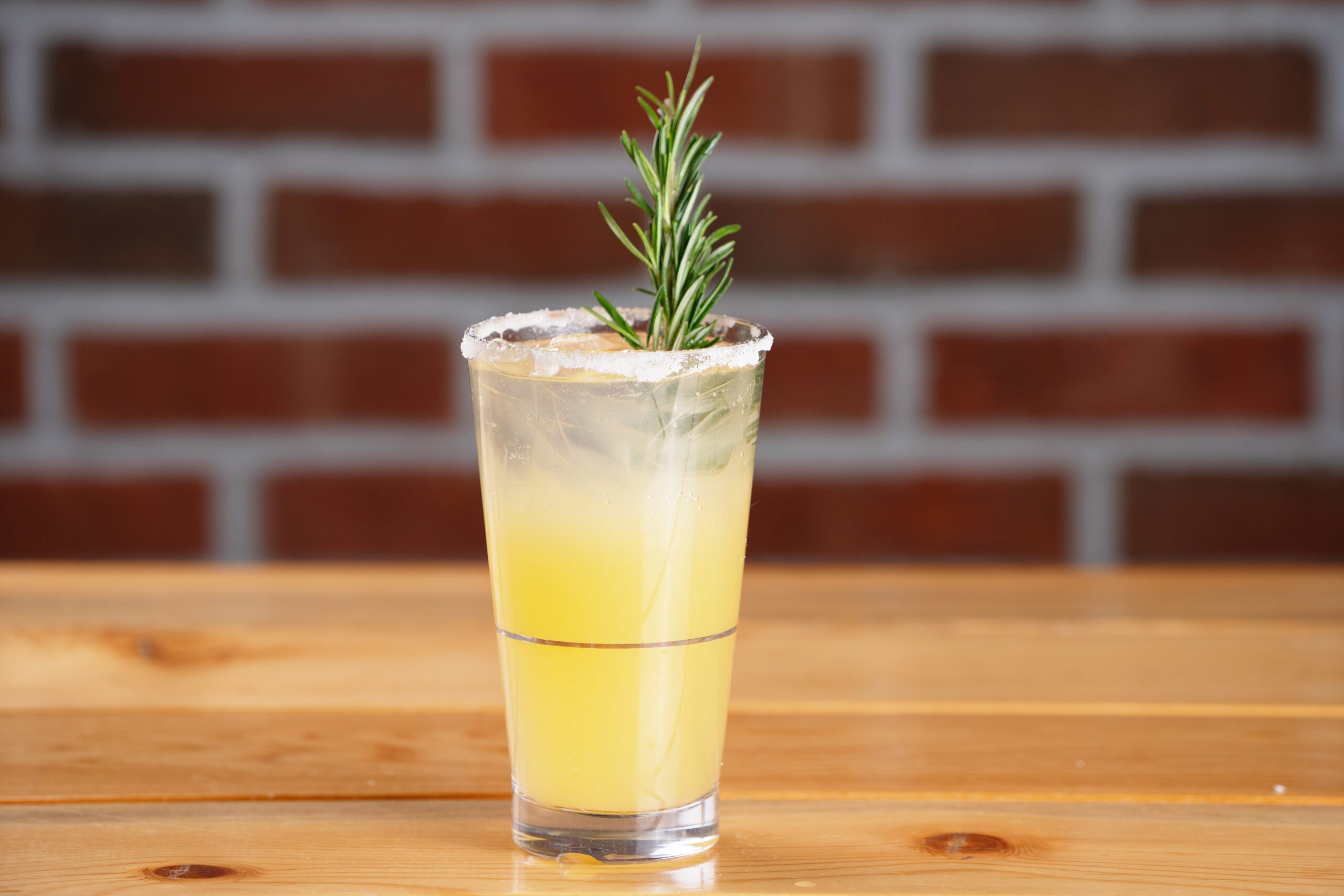 A yellow cocktail in a glass with a salted rim, garnished with a sprig of rosemary, sits on a wooden table with a blurred brick wall in the background.