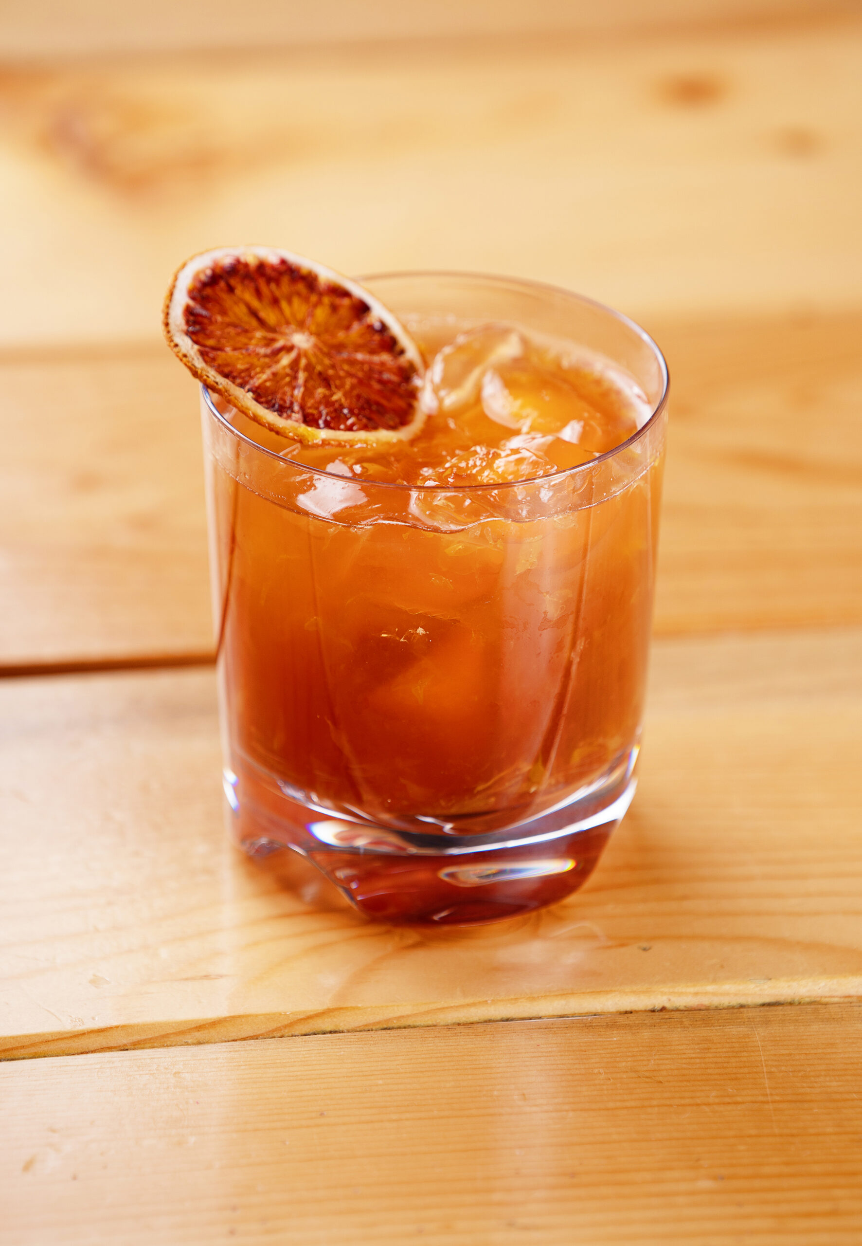 A glass of iced amber-colored cocktail garnished with a dried orange slice sits on a light wooden surface.