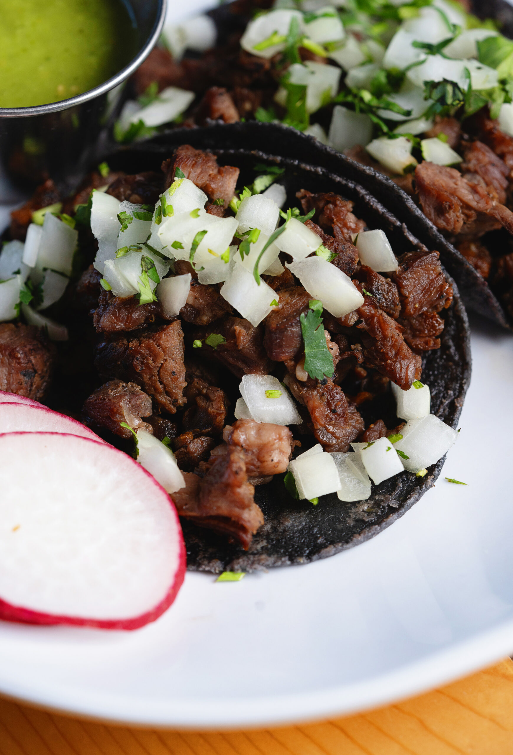 Close-up of two tacos filled with diced grilled beef, chopped white onions, and cilantro on blue corn tortillas, garnished with a radish slice and served with green salsa in a small cup on the side.