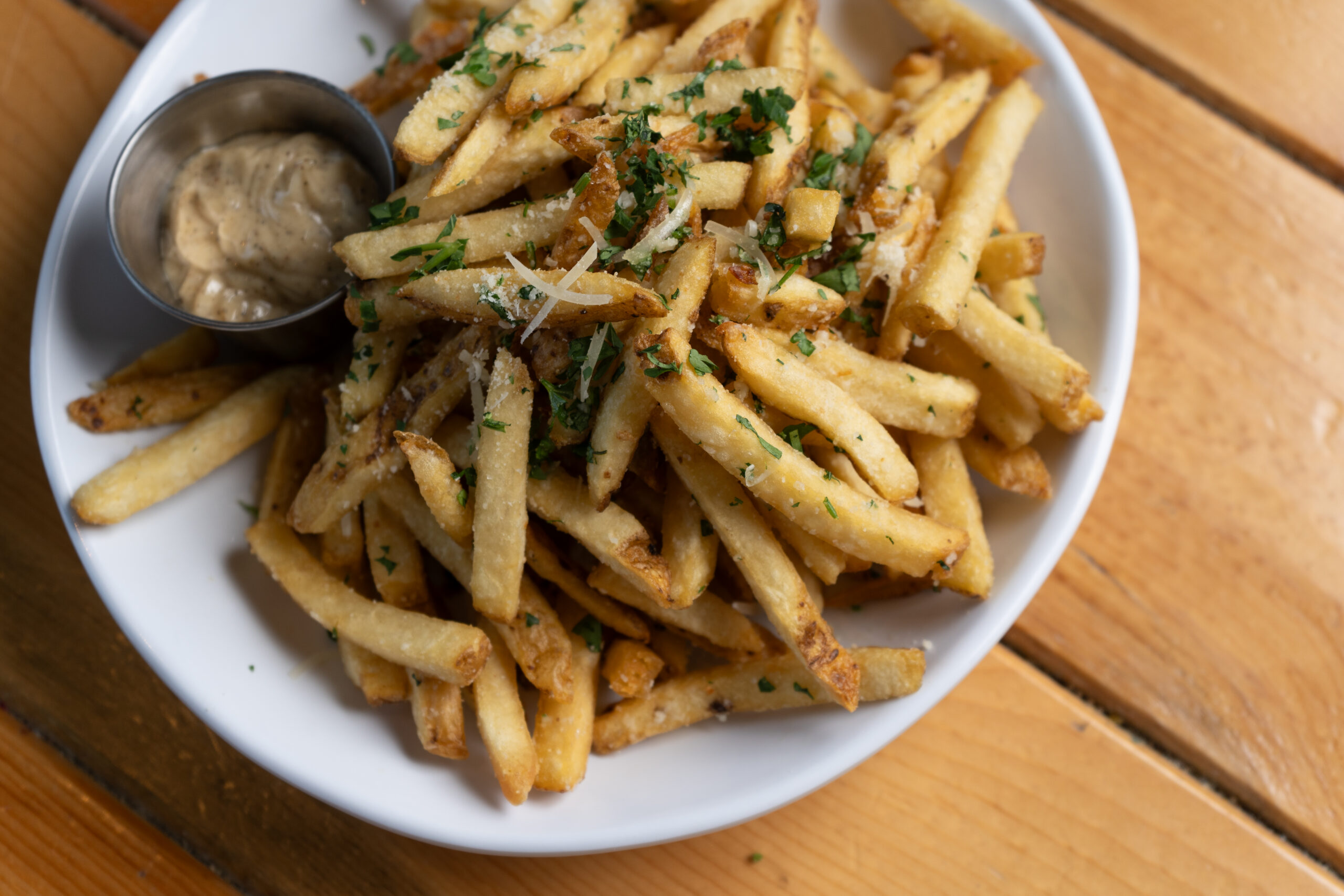 A white plate of seasoned French fries garnished with chopped parsley and shredded cheese, served with a small metal cup of creamy dipping sauce on a wooden table.