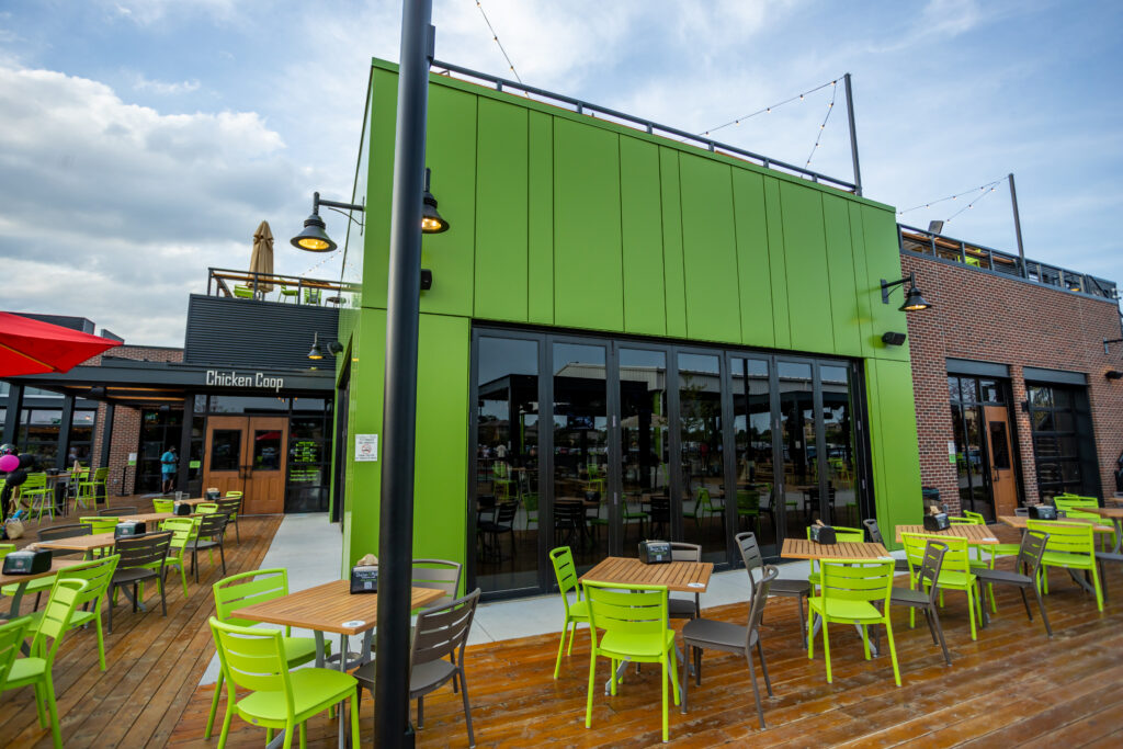 A modern outdoor dining area with green tables and chairs on a wooden deck, in front of a building with bright green walls and large windows. The sky is partly cloudy.