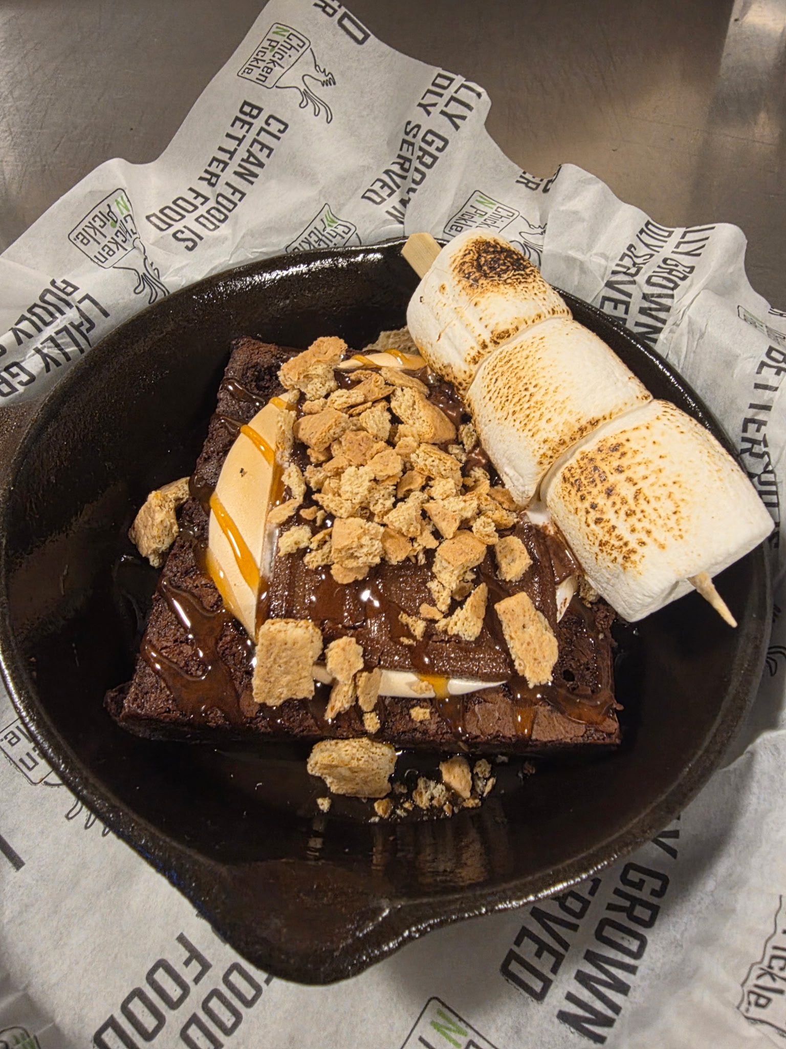 A cast iron skillet with a brownie topped with caramel and chocolate sauce, crushed graham crackers, and a skewer of toasted marshmallows served on branded paper.