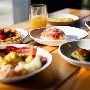 A wooden table set with plates of assorted breakfast foods, including waffles with berries and whipped cream, toast with prosciutto and onions, bacon, eggs, fruit, and a glass of orange juice. Sunlight shines on the meal.