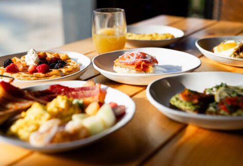 A wooden table set with plates of assorted breakfast foods, including waffles with berries and whipped cream, toast with prosciutto and onions, bacon, eggs, fruit, and a glass of orange juice. Sunlight shines on the meal.