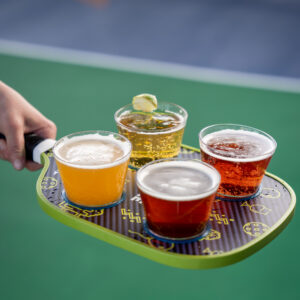 A hand holds a pickleball paddle used as a tray, carrying four plastic cups filled with different types of beer, with a green court surface in the background.