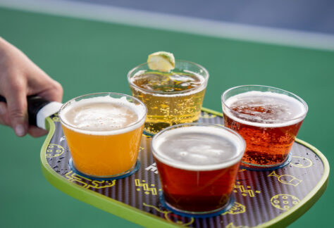 A hand holds a pickleball paddle used as a tray, carrying four plastic cups filled with different types of beer, with a green court surface in the background.