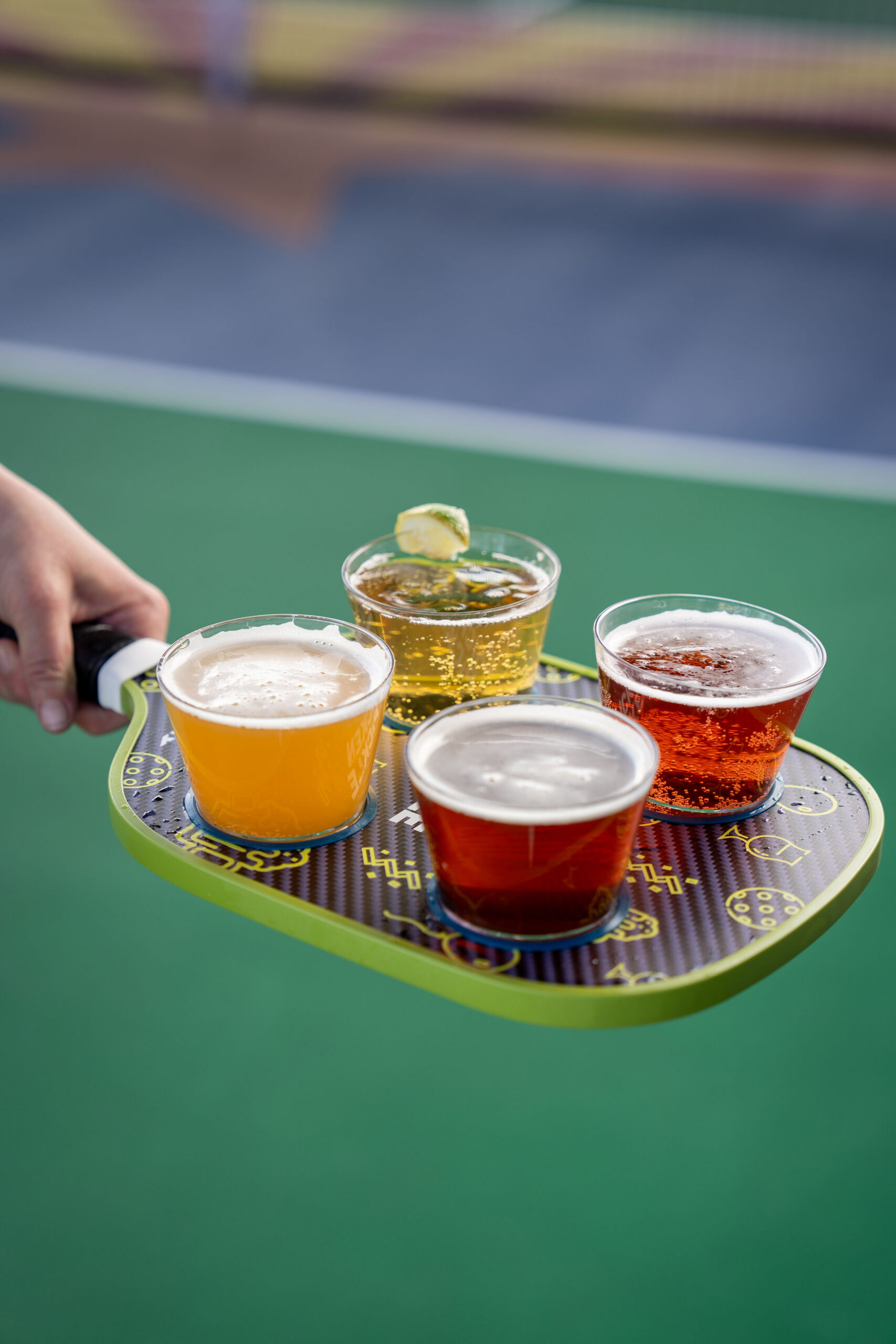A hand holds a pickleball paddle used as a tray, carrying four plastic cups filled with different types of beer, with a green court surface in the background.
