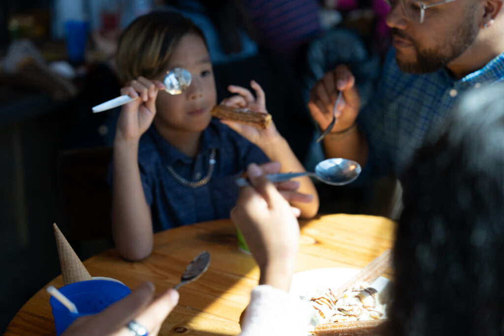 A child holding a spoon up to their face and a dessert bar in the other hand, sitting at a wooden table with three adults, all eating dessert in a warmly lit setting.
