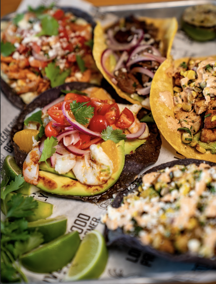 A close-up of colorful gourmet tacos with various toppings, including shrimp, avocado, tomatoes, onions, cilantro, cheese, and grilled corn, served on blue and yellow tortillas with lime wedges on the side.