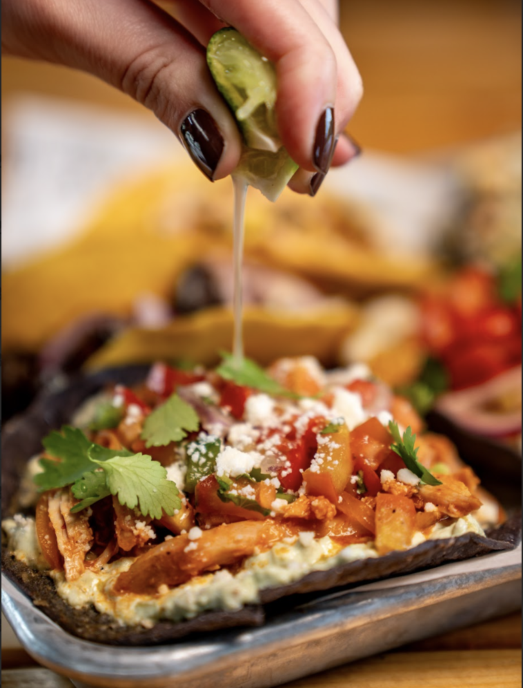 A hand squeezes a lime over a colorful tostada topped with shredded chicken, cheese, tomatoes, onions, and fresh cilantro on a metal tray.