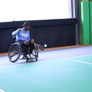 A man in a wheelchair is playing adaptive pickleball at Chicken N Pickle Glendale, preparing to hit the ball with his racket. Another player in a wheelchair is visible in the background on the right side of the court.