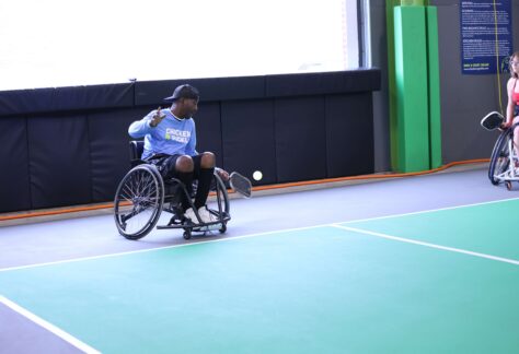 A man in a wheelchair is playing adaptive pickleball at Chicken N Pickle Glendale, preparing to hit the ball with his racket. Another player in a wheelchair is visible in the background on the right side of the court.