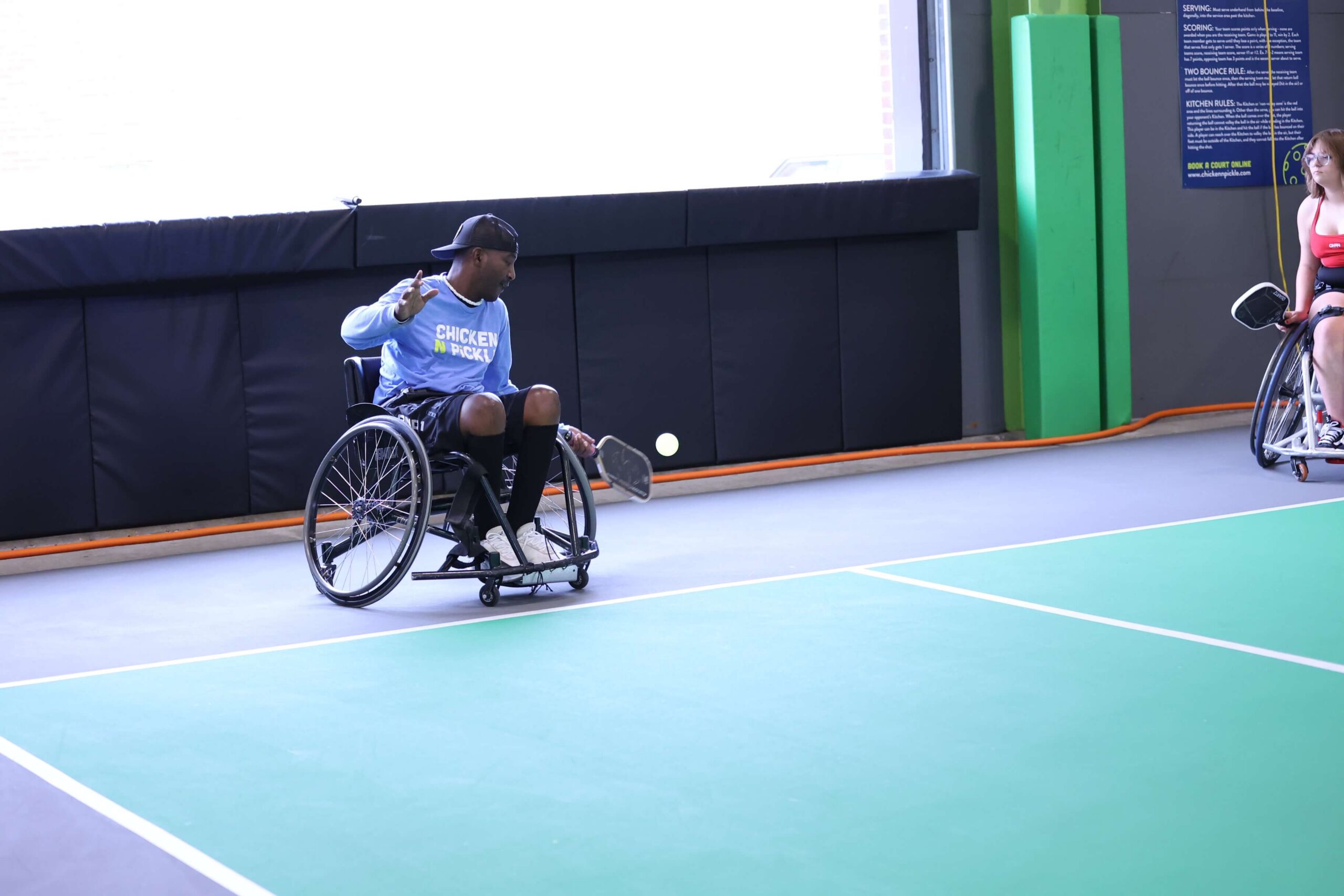 A man in a wheelchair is playing adaptive pickleball at Chicken N Pickle Glendale, preparing to hit the ball with his racket. Another player in a wheelchair is visible in the background on the right side of the court.