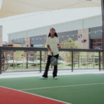A young woman stands on an outdoor pickleball court at Doggy Days at Chicken N Pickle Allen, holding a paddle and appearing ready to play. She is dressed casually, with buildings and a seated person visible in the background.