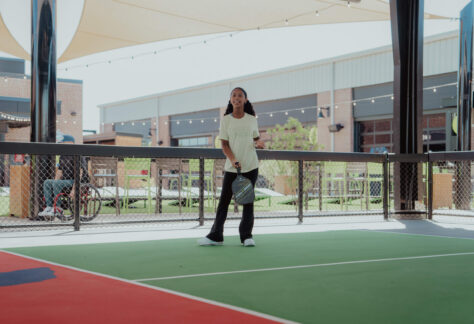 A young woman stands on an outdoor pickleball court at Doggy Days at Chicken N Pickle Allen, holding a paddle and appearing ready to play. She is dressed casually, with buildings and a seated person visible in the background.