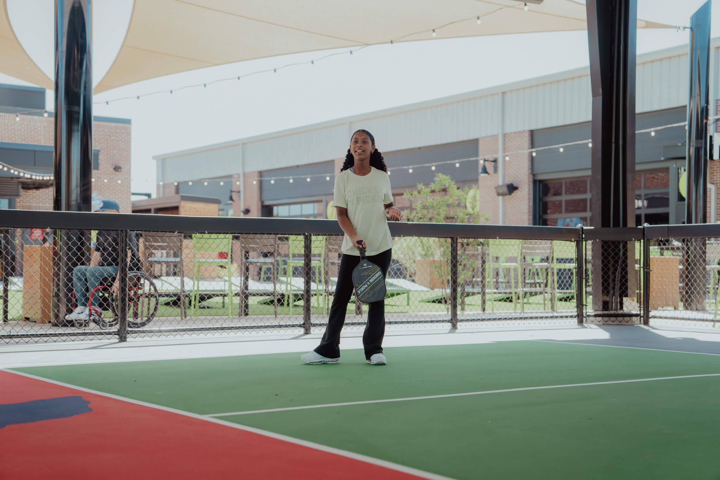 A young woman stands on an outdoor pickleball court at Doggy Days at Chicken N Pickle Allen, holding a paddle and appearing ready to play. She is dressed casually, with buildings and a seated person visible in the background.