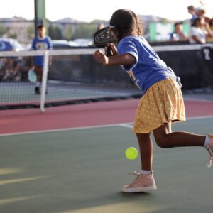 A young girl wearing a blue shirt, yellow polka dot skirt, and pink high-top sneakers plays pickleball outdoors, swinging her paddle at a yellow ball, with spectators watching in the background.