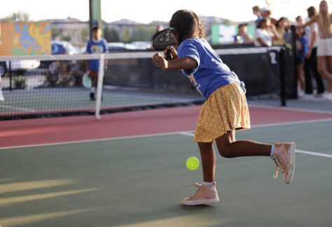 A young girl wearing a blue shirt, yellow polka dot skirt, and pink high-top sneakers plays pickleball outdoors, swinging her paddle at a yellow ball, with spectators watching in the background.