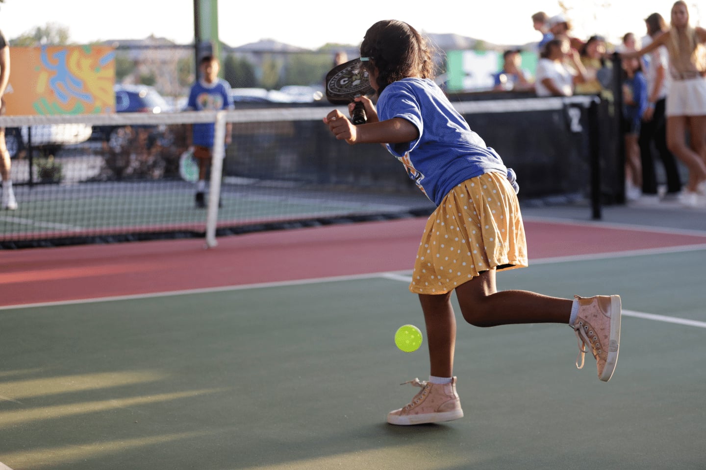 A young girl wearing a blue shirt, yellow polka dot skirt, and pink high-top sneakers plays pickleball outdoors, swinging her paddle at a yellow ball, with spectators watching in the background.