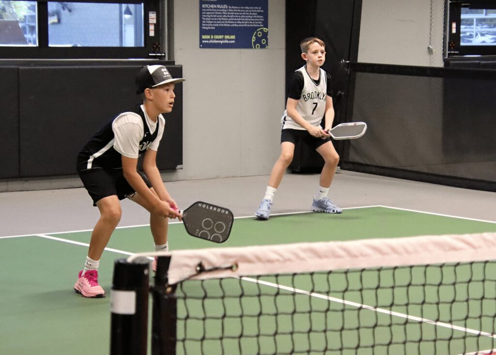 Two boys in sports uniforms and sneakers stand ready with paddles on an indoor pickleball court, preparing to receive a serve. A net runs across the foreground.