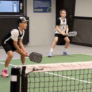 Two boys in sports uniforms and sneakers stand ready with paddles on an indoor pickleball court, preparing to receive a serve. A net runs across the foreground.