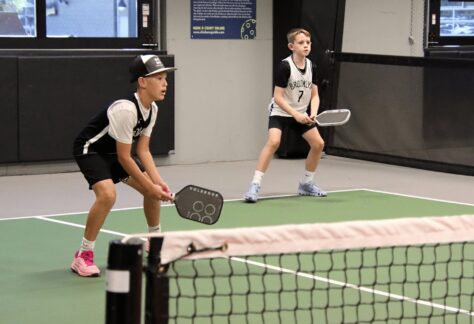 Two boys in sports uniforms and sneakers stand ready with paddles on an indoor pickleball court, preparing to receive a serve. A net runs across the foreground.