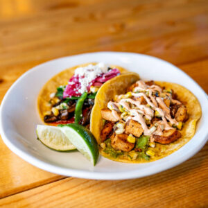 A white plate with two tacos—one with grilled vegetables and cheese, the other with seasoned chicken, corn, and sauce—sits on a wooden table at Chicken N Pickle Kansas City. A lime wedge adds zest to this Taco Tuesday delight.