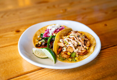 A white plate with two tacos—one with grilled vegetables and cheese, the other with seasoned chicken, corn, and sauce—sits on a wooden table at Chicken N Pickle Kansas City. A lime wedge adds zest to this Taco Tuesday delight.