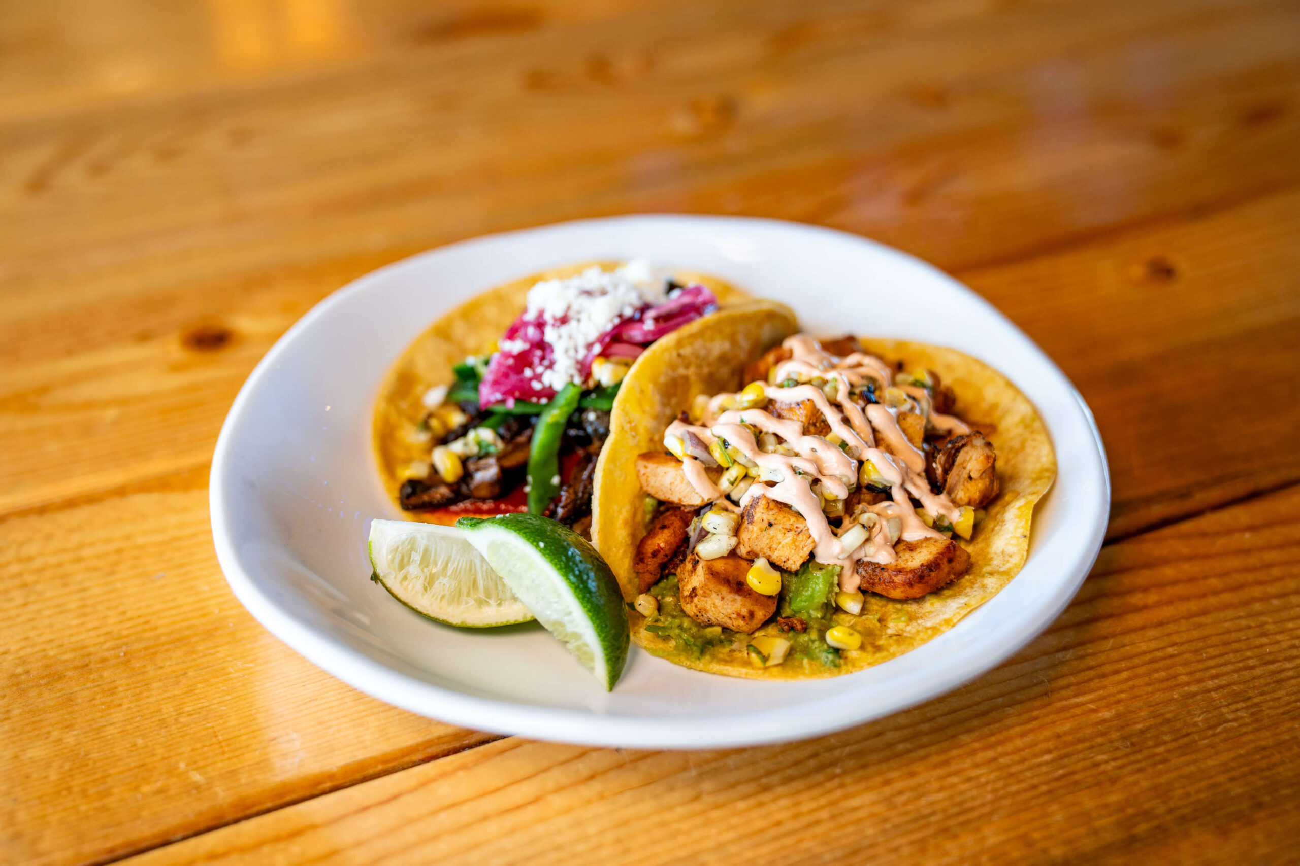 A white plate with two tacos—one with grilled vegetables and cheese, the other with seasoned chicken, corn, and sauce—sits on a wooden table at Chicken N Pickle Kansas City. A lime wedge adds zest to this Taco Tuesday delight.