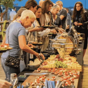 A group of people serves themselves food from a buffet table outdoors. The table is filled with appetizers, fruits, cheeses, and various plated dishes. The setting appears casual and social, with people chatting and helping themselves.