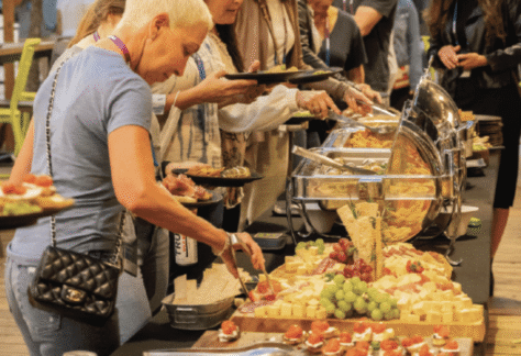A group of people serves themselves food from a buffet table outdoors. The table is filled with appetizers, fruits, cheeses, and various plated dishes. The setting appears casual and social, with people chatting and helping themselves.