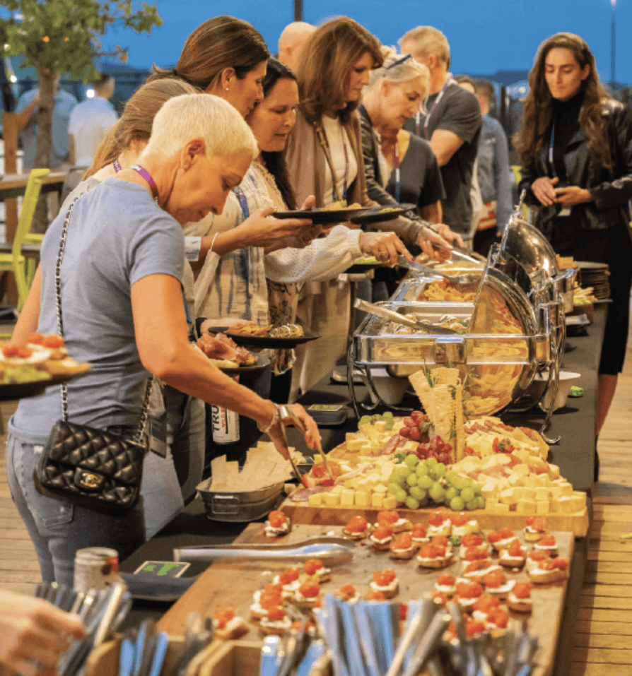 A group of people serves themselves food from a buffet table outdoors. The table is filled with appetizers, fruits, cheeses, and various plated dishes. The setting appears casual and social, with people chatting and helping themselves.