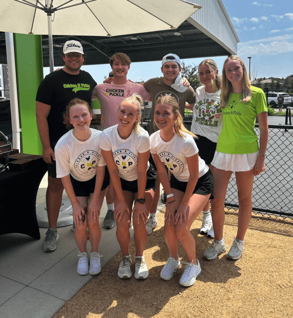 Eight young adults, four men in the back row and four women in the front row, smile for a group photo outdoors at a pickleball event. Some wear Chicken N Pickle shirts and athletic clothing.
