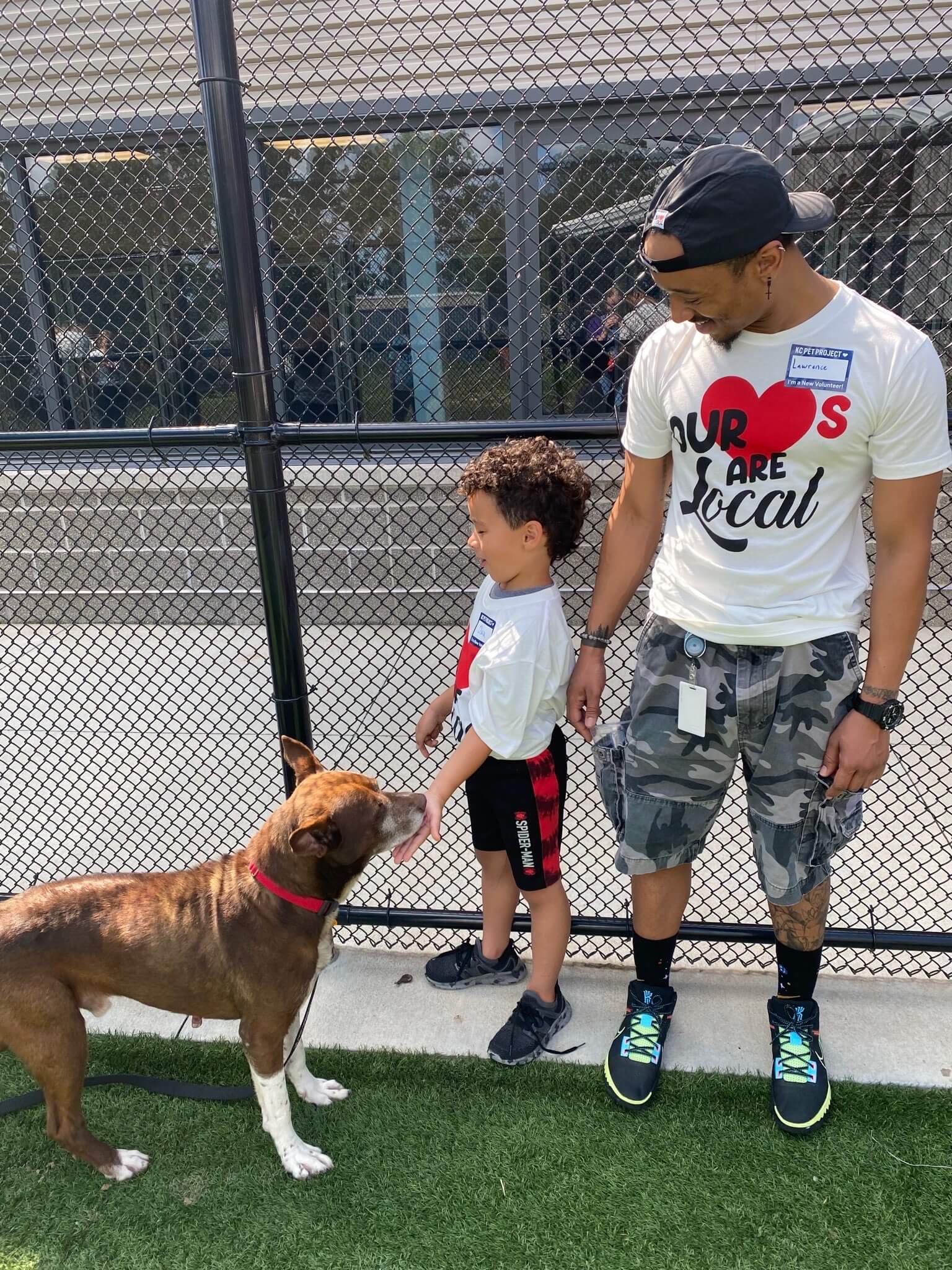 A man and a young boy stand on grass by a chain-link fence, holding hands. The boy is petting a brown and white dog. Both wear white shirts; the man also wears a cap and camo shorts.
