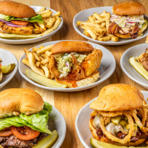 Seven plates of assorted burgers with lettuce, tomato, onions, cheese, and various toppings, each served with French fries and a pickle spear, arranged on a wooden table.