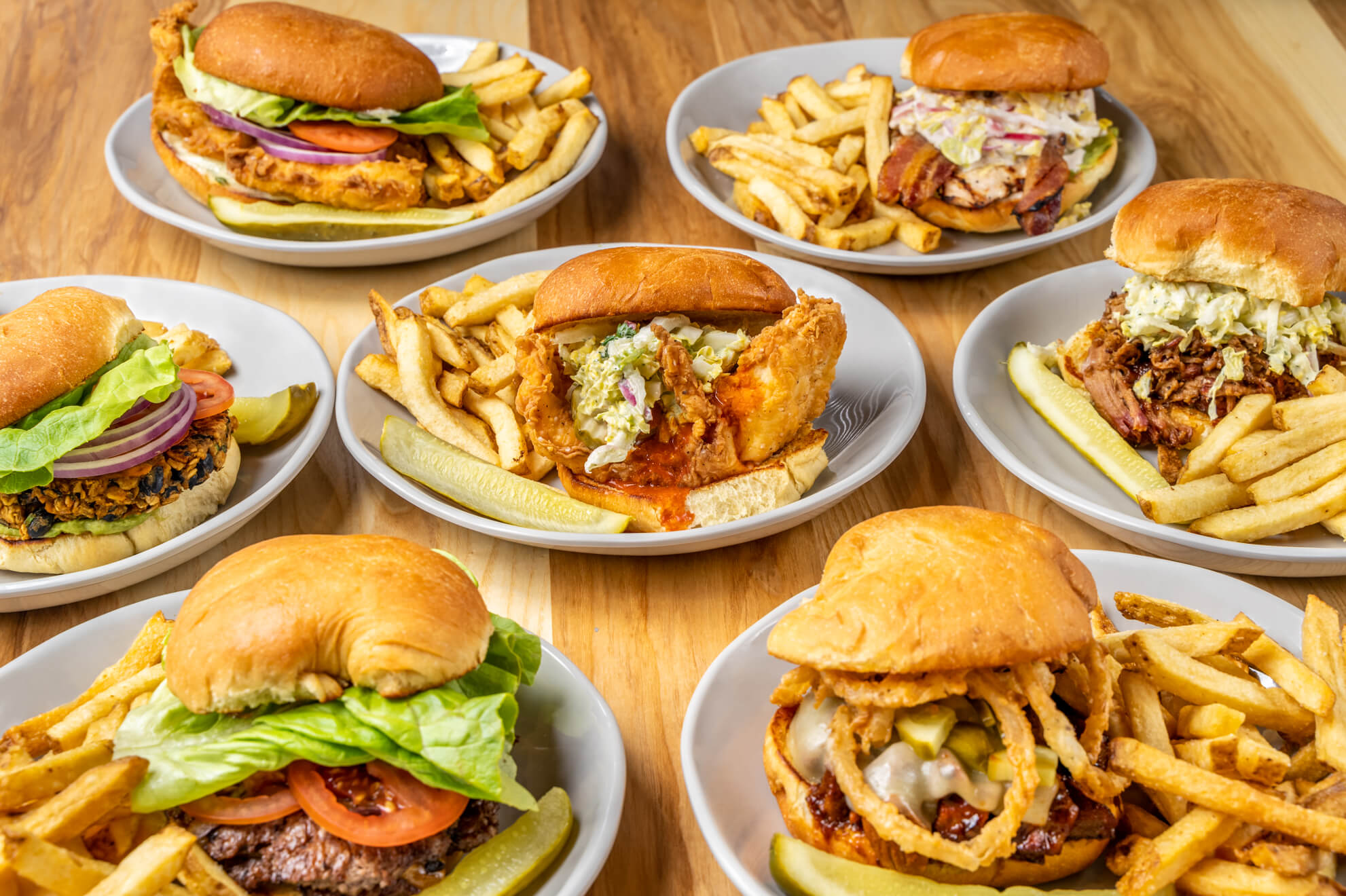 Seven plates of assorted burgers with lettuce, tomato, onions, cheese, and various toppings, each served with French fries and a pickle spear, arranged on a wooden table.