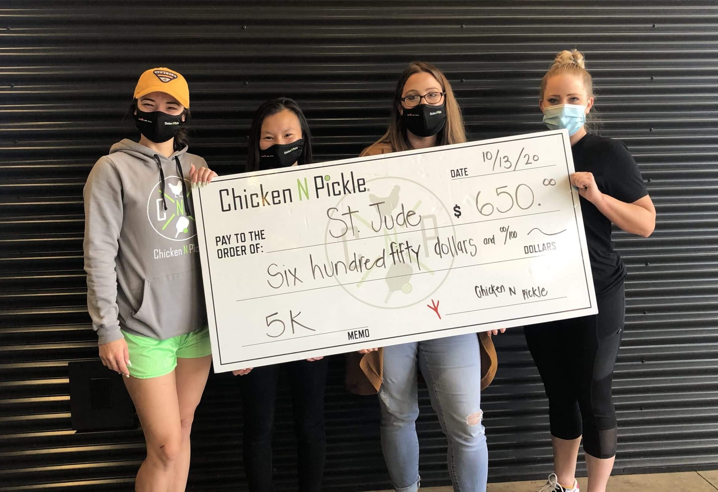 Four women stand in front of a striped wall, smiling and holding an oversized check for $650 made out to St. Jude from Chicken N Pickle, dated 10/13/20, for a 5K fundraiser.
