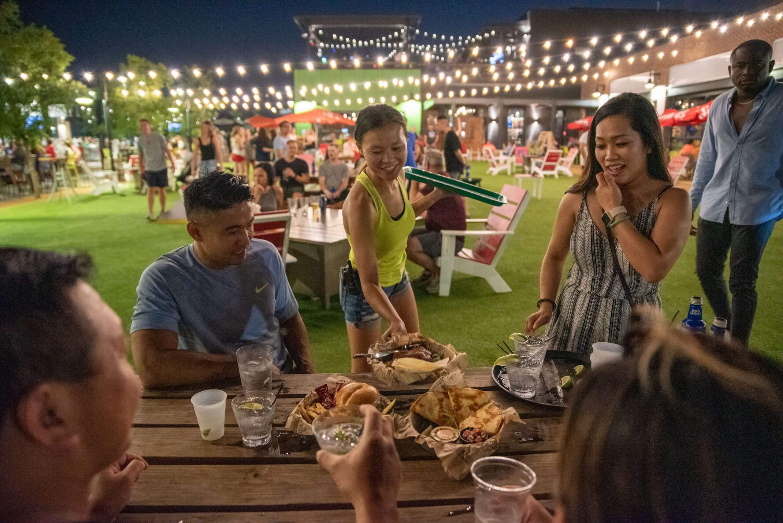 A group of people enjoy food and drinks around a wooden table at an outdoor venue with string lights, green turf, and other groups socializing in the background at night.